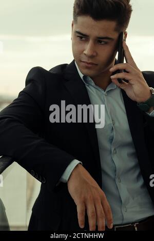 Male entrepreneur in formal suit leaning on metal railing in city and talking on smartphone while looking away Stock Photo