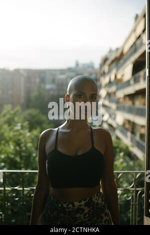 Black female with bald hairstyle and in trendy outfit standing on terrace of outdoors cafe and relaxing while looking at camera Stock Photo
