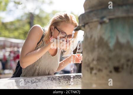 Thirsty young casual cucasian woman wearing medical face mask drinking water from public city fountain on a hot summer day. New social norms during