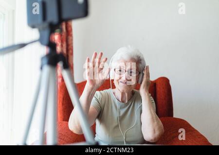 Senior lady in headphones sitting in cozy armchair at home and having video call Stock Photo