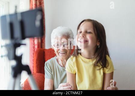 Smiling senior Woman and girl sitting in living room together and having video call on smartphone Stock Photo