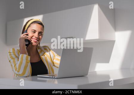 Modern female freelancer in stylish outfit sitting at counter with laptop and discussing details of project via mobile phone while working in contemporary workspace Stock Photo