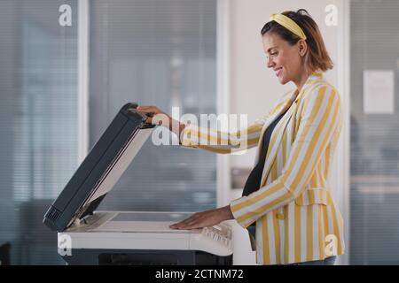 Side view of optimistic pregnant businessWoman in stylish outfit putting paper sheet on photocopier while working in modern workspace Stock Photo