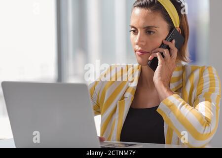 Modern female freelancer in stylish outfit sitting at counter with laptop and discussing details of project via mobile phone while working in contemporary workspace Stock Photo
