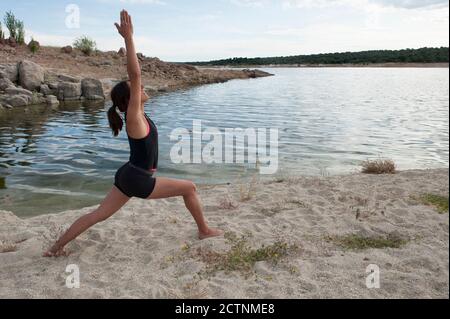 Full body side view of slim female in sportswear practicing Warrior asana with arms raised while doing yoga near lake Stock Photo