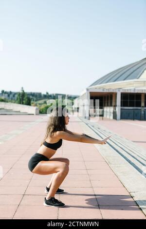 Full length side view of motivated strong fit female in sportswear doing squad exercise during intense training on paved square in city Stock Photo