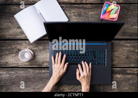 Top view of female hands typing on laptop computer with candle, colorful markers and open notepad beside it on a rustic wooden desk. Stock Photo