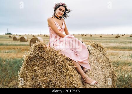 Young brunette in a pink satin dress sitting on a hay bale. A woman has a wreath on her head and holds a bouquet of wildflowers in her hands. Stock Photo