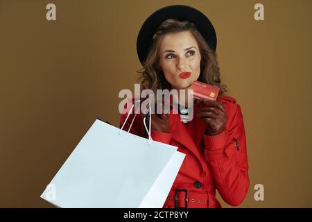 Hello autumn. pensive trendy housewife in red coat and black beret with credit card, leather gloves and paper shopping bags against bronze background. Stock Photo