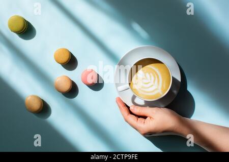 Morning coffee in a woman's hand with delicious macaroons scattered around in the sunlight.