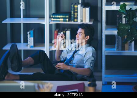 Thai’s man relaxed confident young businessman sleep with legs on desk at office work late overtime at night Table footrest Stock Photo