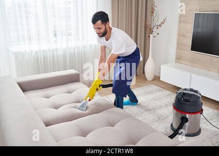 Close-up of housekeeper holding modern washing vacuum cleaner and cleaning dirty sofa with professionally detergent. Professional springclean at home Stock Photo