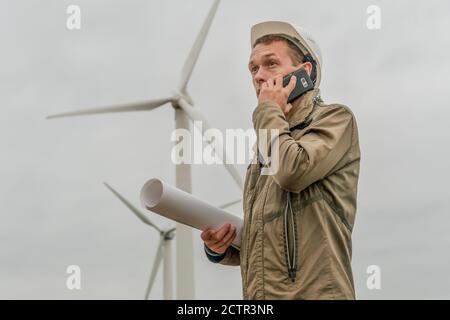 Engineer with a plan in hand talking on a mobile phone near wind generators in cloudy weather Stock Photo