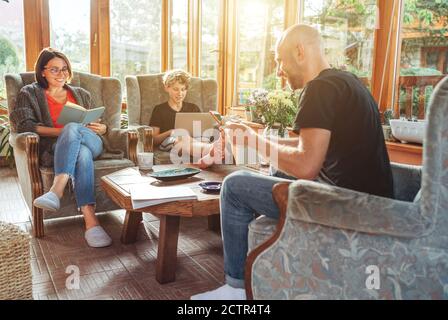 Mother, father and son family sitting together at Sunroom in cozy armchairs and reading books, using laptop or browsing a smartphone. Stock Photo