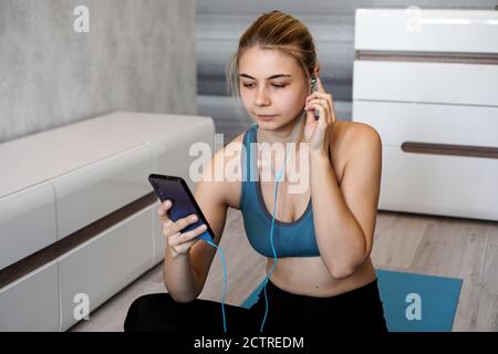 Take sport and add music. Female athlete with earphones enjoying the music playing and touching a screen of her phone while sitting on the floor. Stock Photo