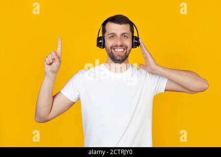 Young man with a beard in a white t-shirt listens to music with headphones over isolated yellow background, surprised with an idea or question, pointing with a finger with a happy face, number one. Stock Photo