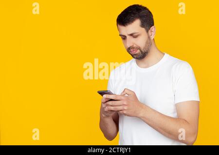 A young man with a beard in a white t-shirt uses a smartphone with a confident expression on his smart face, thinking seriously. Stands on isolated yellow background. Stock Photo