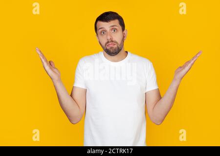 A young man with a beard in a white t-shirt is an ignorant and confused expression with raised arms and hands. Doubt concept. Stands on isolated yellow background. Stock Photo
