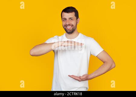 Young man with a beard in a white T-shirt gesturing with his hands showing a large and large size sign, a symbol of measure. Smiling looking into the camera. Stands on isolated yellow background. Stock Photo