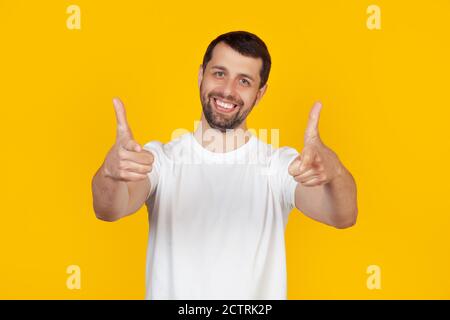 Young man with a beard in a white t-shirt pointing with his fingers at the camera with a happy and funny face. Good energy and vibes. Stands on isolated yellow background. Stock Photo