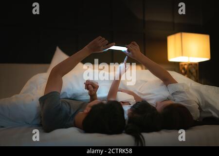 Asian happy family mother, father and little daughter watching movie or cartoon in smartphone together and blanket cover their head in bed at night at