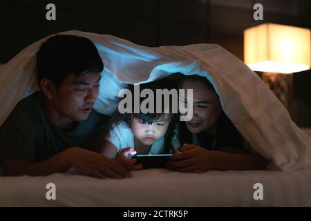 Asian happy family mother, father and little daughter watching movie or cartoon in smartphone together and blanket cover their head in bed at night at Stock Photo