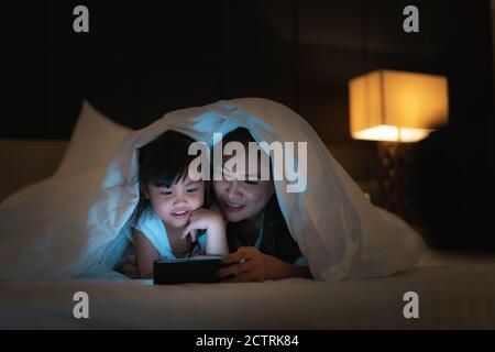 Asian happy family mother and little daughter watching movie or cartoon in smartphone together and blanket cover their head in bed at night at home