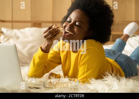 Happy Afro-American woman in yellow sweater relaxing, eating popcorn while watching movie on laptop, talking on video chat, lying in bed. Lazy rest at Stock Photo