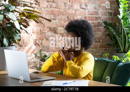 Side view smiling Afro-American biracial millennial woman wearing headset, communicating via video call on laptop, talking in zoom app, watching webin Stock Photo