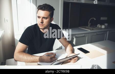 Concentrated caucasian man with fashionable haircut is working remotely from home and using tablet while write something in the kitchen Stock Photo