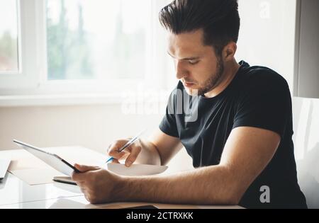 Bearded caucasian businessman working from home using a tablet and some papers wearing a black t-shirt in the living room Stock Photo