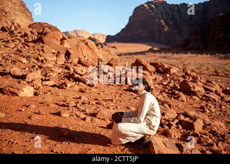 Wadi Rum, Jordan 03/31/2010: A young bedouin man wearing long white jubba thobe dress and sandals is sitting on a rock in Wadi Rum Desert. Sandstone c Stock Photo