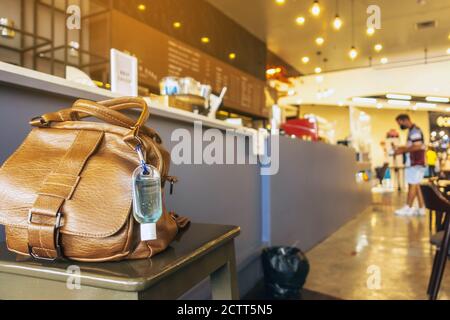 Mini portable alcohol gel bottle to kill Corona Virus(Covid-19) hang on a brown leather shoulder bag on chair in coffee shop.New normal lifestyle. Hea Stock Photo