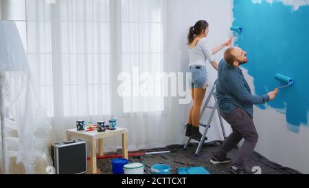 Cheerful couple painting and dancing while redecorating living room. Happy family. Apartment redecoration and home construction while renovating and improving. Repair and decorating. Stock Photo