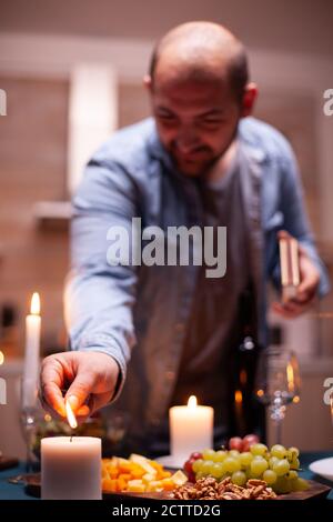 Husband lighting candle on kitchen table a for festive dinner with wife. Man preparing festive meal with healthy food for anniversary celebration, romantic date. Stock Photo