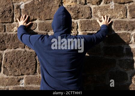 Back view of a man in blue hoodie, facing stone wall, hands up spread on the wall Stock Photo
