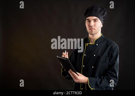 Portrait of young male dressed in a black chef suit takes notes, looking at the camera posing on a brown background with copy space advertising area