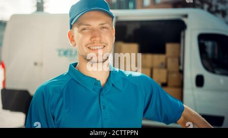 Portrait of Handsome Delivery Man Holds Cardboard Box Package Standing in Modern Stylish Business District with Delivery Van in Background. Smiling Stock Photo