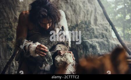 Close-up Shot of a Primeval Caveman Wearing Animal Skin Hits Rock with Sharp Stone, Makes First Primitive Tool for Hunting Animal Prey. Neanderthal Stock Photo