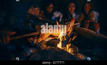 Neanderthal or Homo Sapiens Family Cooking Animal Meat over Bonfire and then Eating it. Tribe of Prehistoric Hunter-Gatherers Wearing Animal Skins Stock Photo