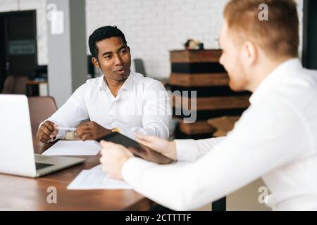 Two young multiethnic businessmen sitting together at office desk and having business team meeting Stock Photo