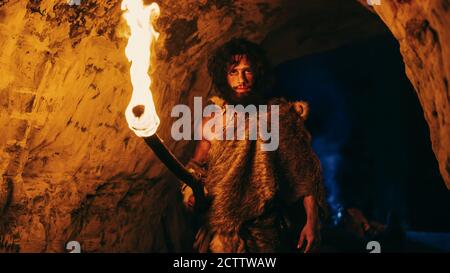 Portrait of Primeval Caveman Wearing Animal Skin Exploring Cave At Night, Holding Torch with Fire Looking into Camera at Night. Stock Photo