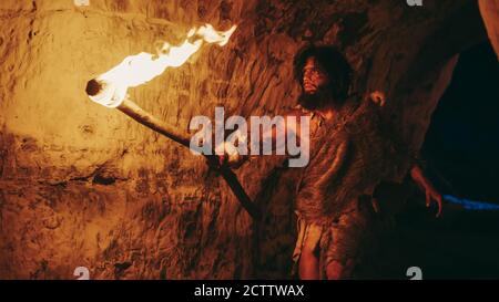 Primeval Caveman Wearing Animal Skin Exploring Cave At Night, Holding Torch with Fire Looking at Drawings on the Walls at Night. Neanderthal Searching Stock Photo