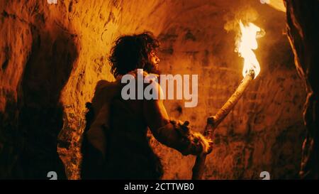 Portrait of Primeval Caveman Wearing Animal Skin Exploring Cave At Night, Holding Torch with Fire Looking at Drawings on the Walls at Night Stock Photo