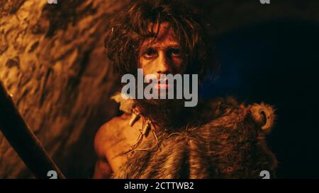 Portrait of Primeval Caveman Wearing Animal Skin Exploring Cave At Night, Holding Torch with Fire Looking into Camera at Night. Stock Photo