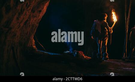Primeval Caveman Wearing Animal Skin Stands in a Cave At Night, Holding Torch with Fire Looking Out of The Cave at Night. Back View Stock Photo