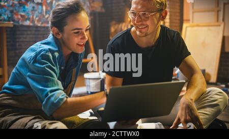 Couple of Artists Sitting in their Studio Use Laptop Computer, Working on a Project, Talking and Smiling. Painter and Her Partner Doing Research Stock Photo
