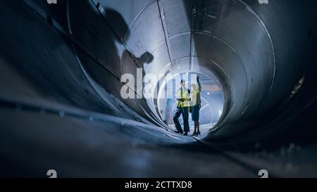 Two Heavy Industry Engineers Walking Inside Pipe, Use Laptop, Have Discussion, Checking Design. Construction of the Oil, Natural Gas and Biofuels Stock Photo