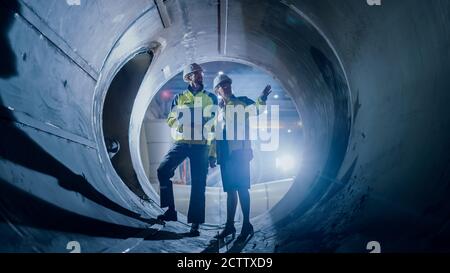Two Heavy Industry Engineers Walking Inside Pipe, Use Laptop, Have Discussion, Checking Design. Construction of the Oil, Natural Gas and Biofuels Stock Photo