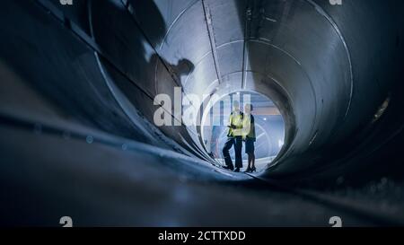 Two Heavy Industry Engineers Walking Inside Pipe, Use Laptop, Have Discussion, Checking Design. Construction of the Oil, Natural Gas and Biofuels Stock Photo
